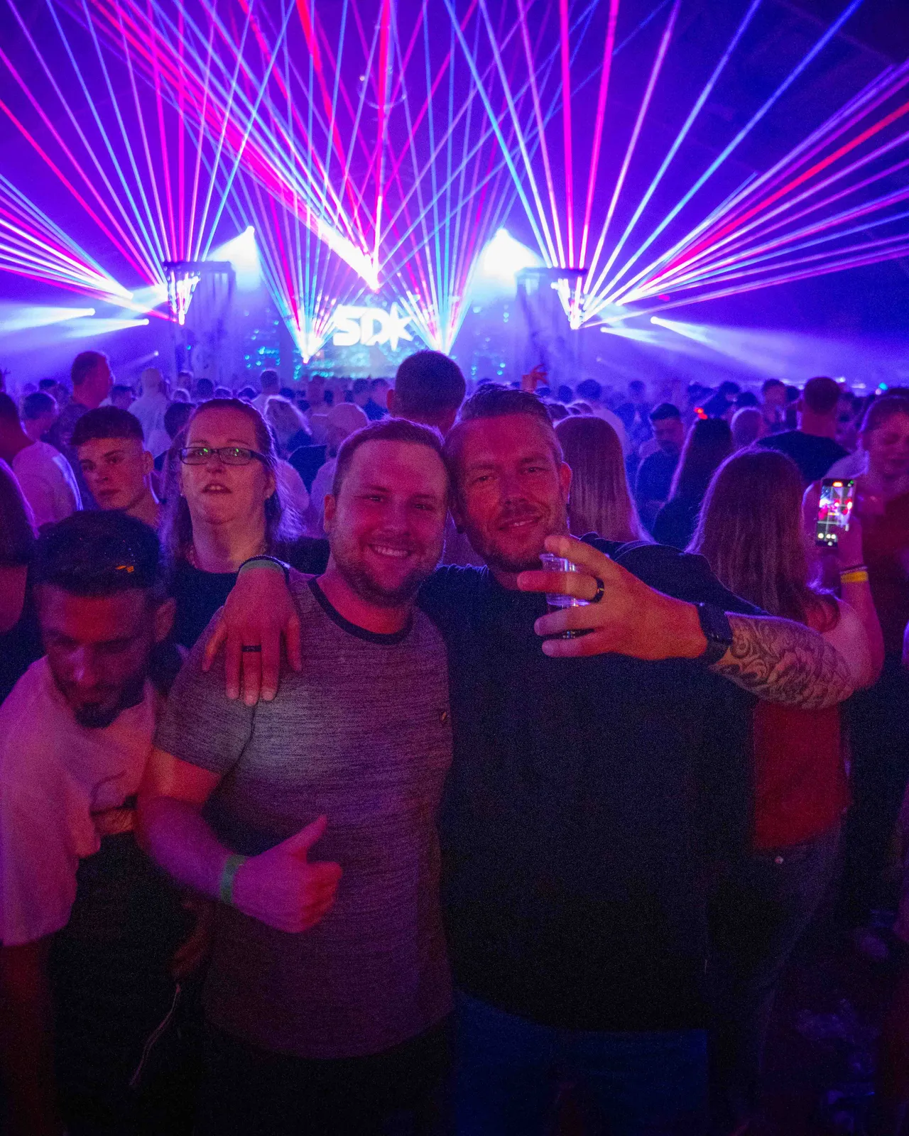 Zwei Gäste mit Rücken zur Bühne und Laser im Hintergrund gucken in Kamera.